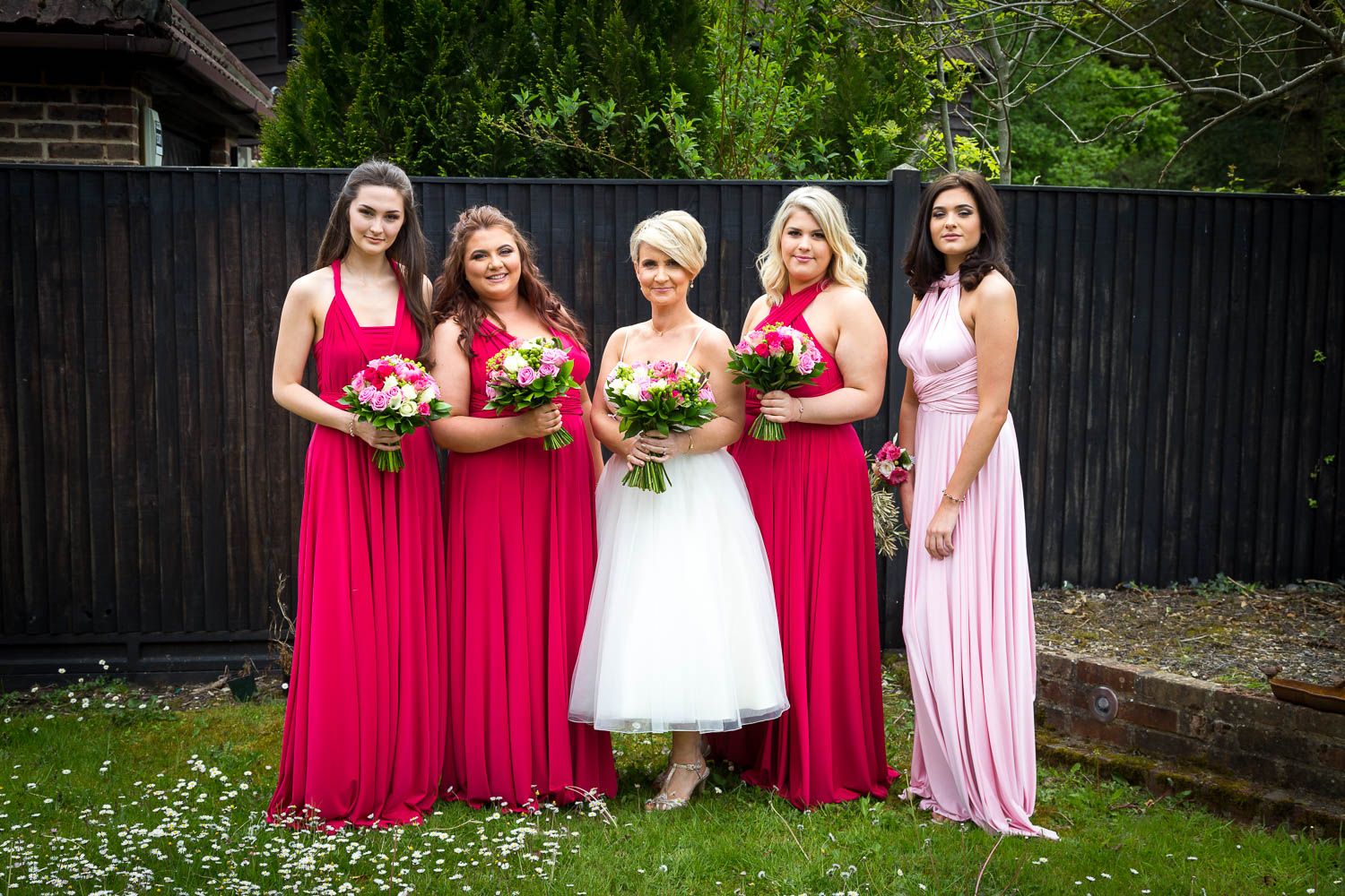 A wedding party poses outdoors with bright pink bridesmaid dresses and bouquets against a dark fence background.