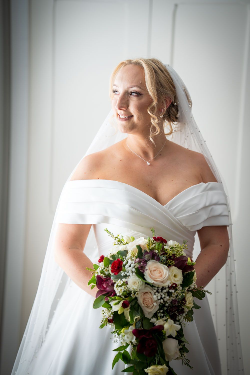 A bride in an elegant off-shoulder white wedding gown holds a beautiful bouquet of burgundy and blush roses.