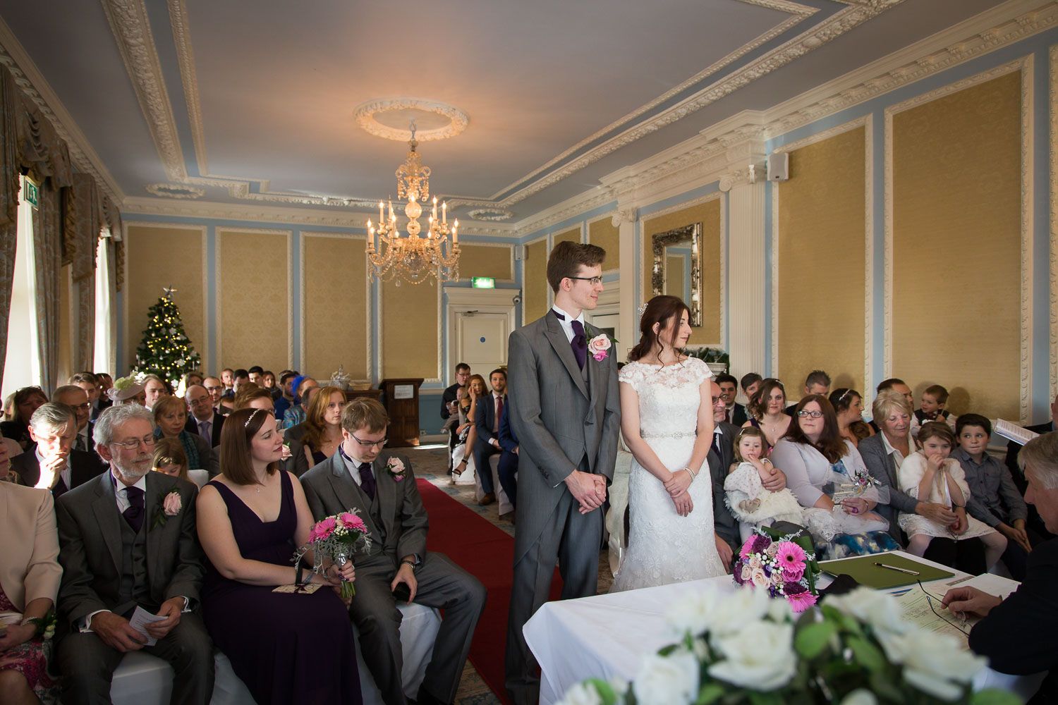An elegant wedding ceremony takes place in a grand ballroom with ornate walls, chandelier and guests seated in rows.