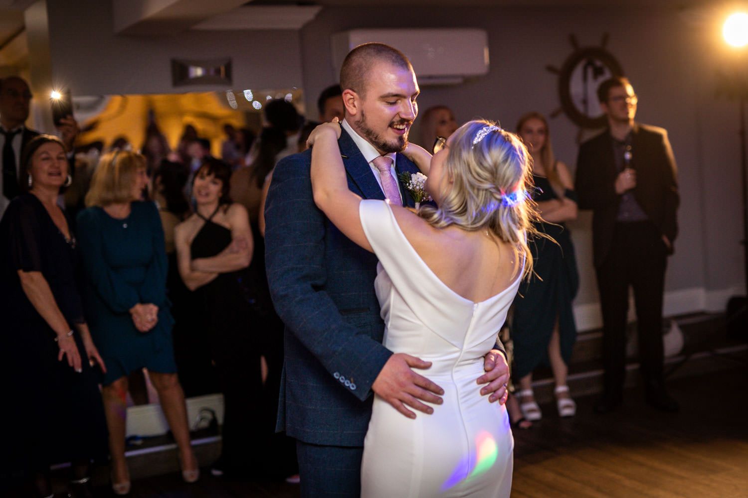 A couple embraces on the dance floor during their wedding reception with colorful party lights in the background.