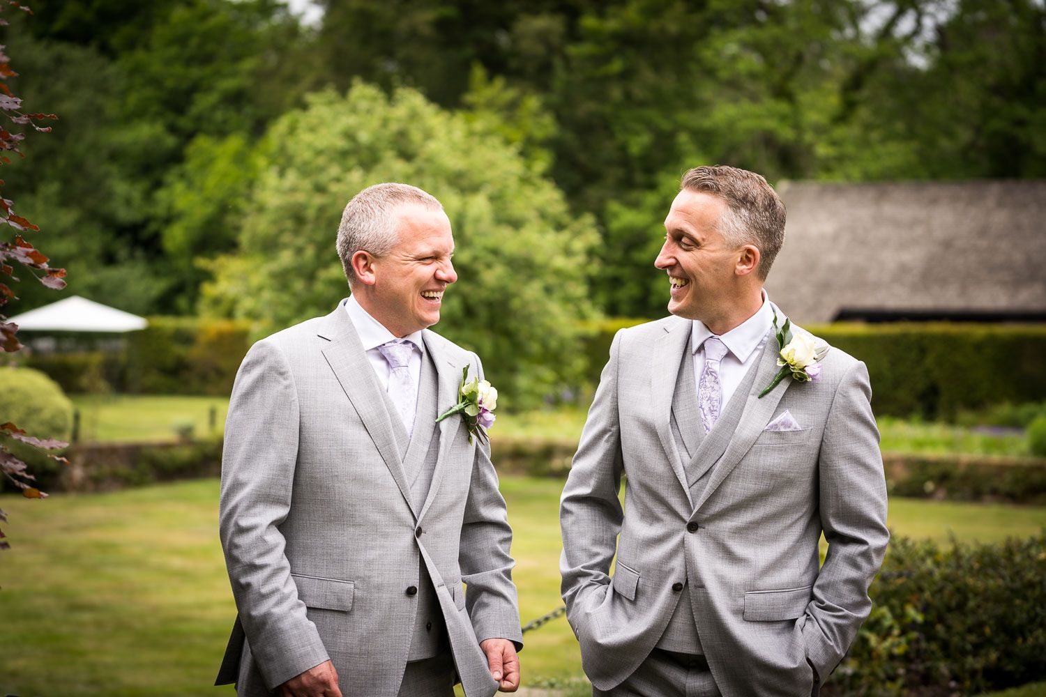 Two men in gray suits and ties share a happy moment together in a lush garden setting.