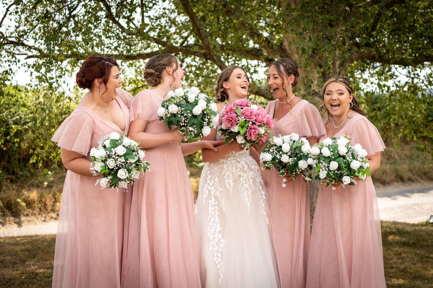 Bridal party wearing blush pink dresses stand together outdoors holding white and pink floral bouquets.