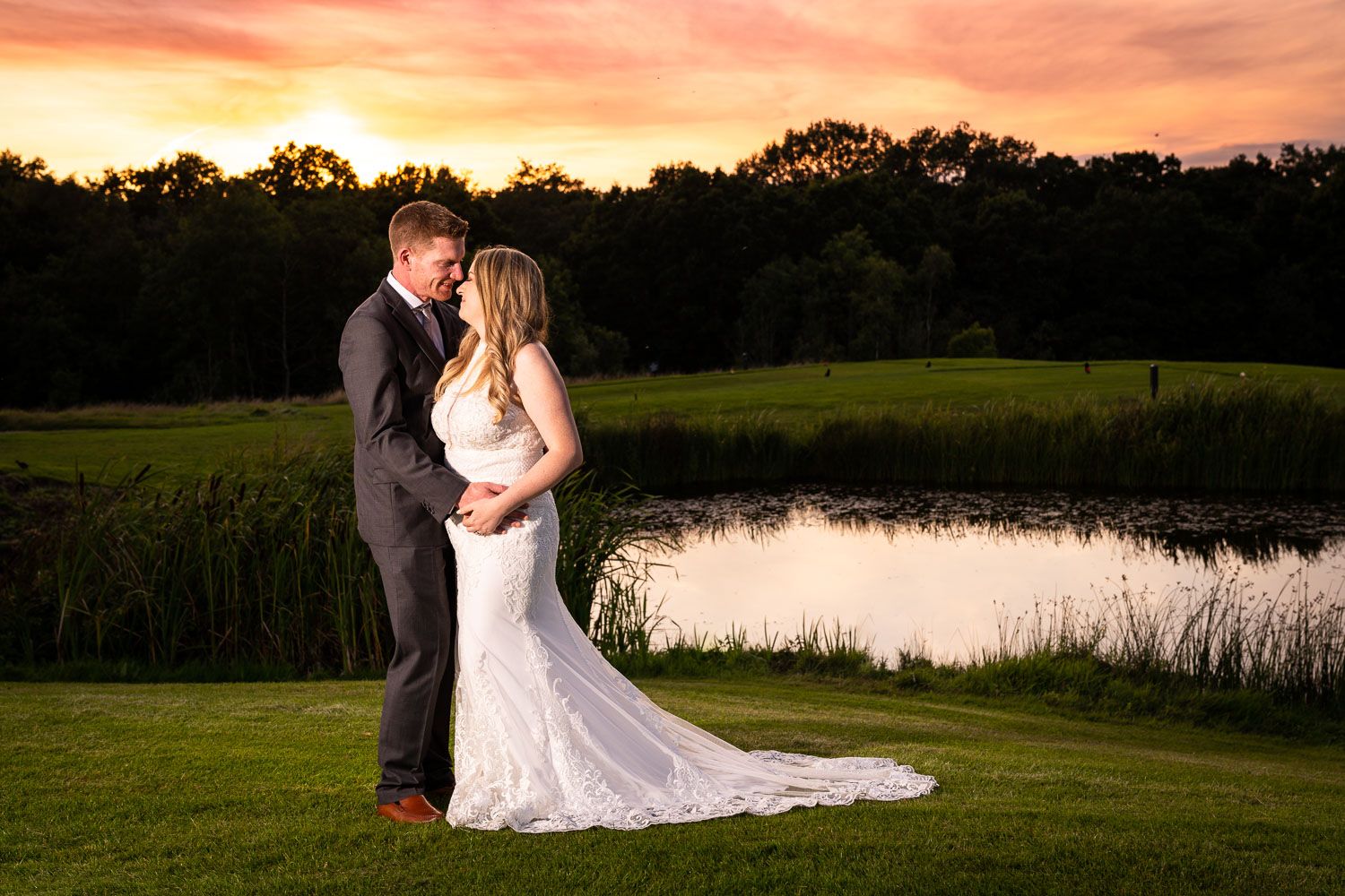 A couple embraces on a golf course at sunset with a pond and trees in the background during their wedding photoshoot.