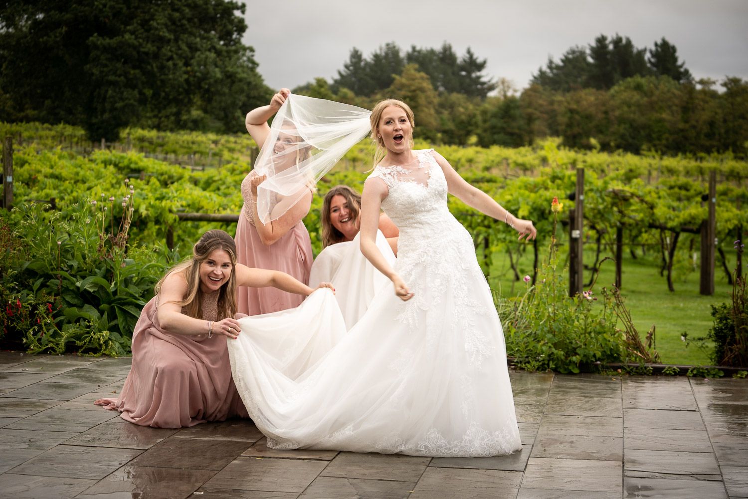 A wedding party poses together on a stone patio surrounded by lush green vineyard scenery.