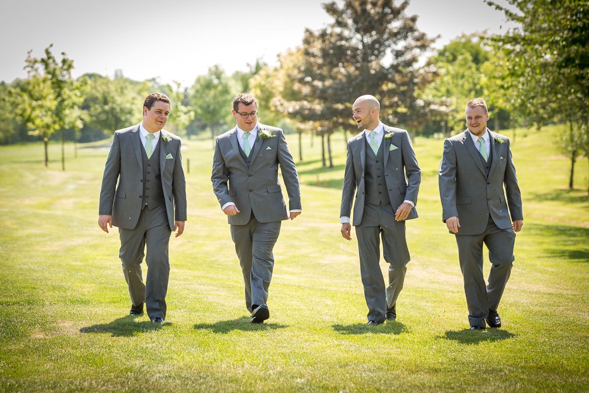 Four groomsmen in matching gray suits walk across a sunlit lawn on a wedding day.