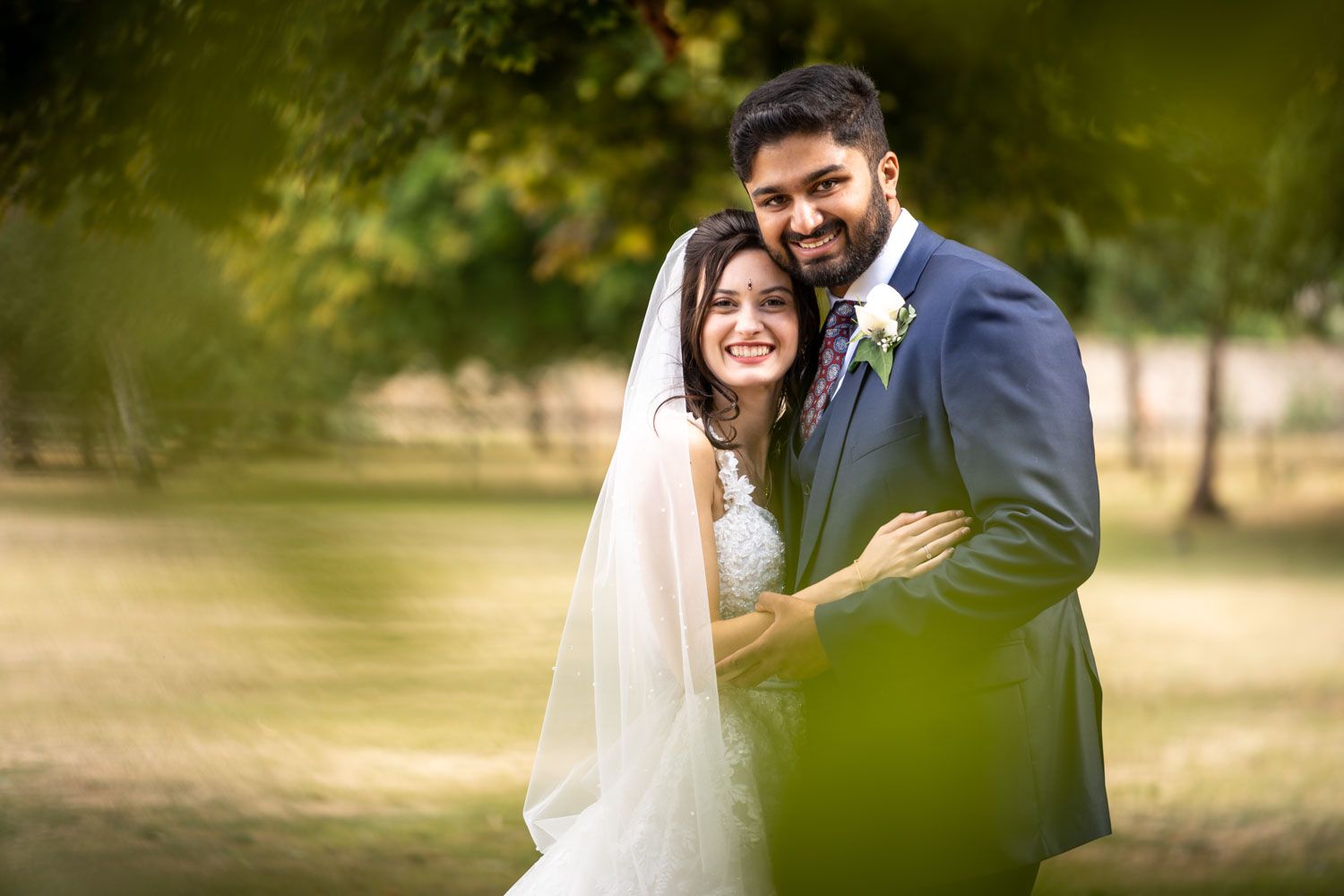 A bride in white dress and groom in navy suit embrace during outdoor wedding photoshoot with soft natural lighting.