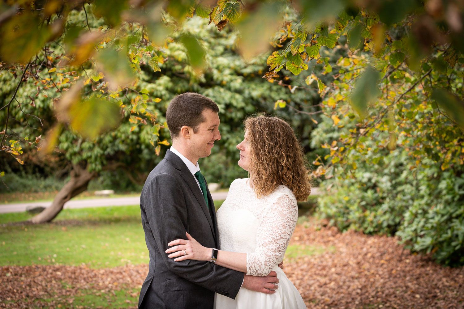 A couple embraces in an outdoor garden setting with autumn foliage and mulch-covered ground during a wedding photoshoot.
