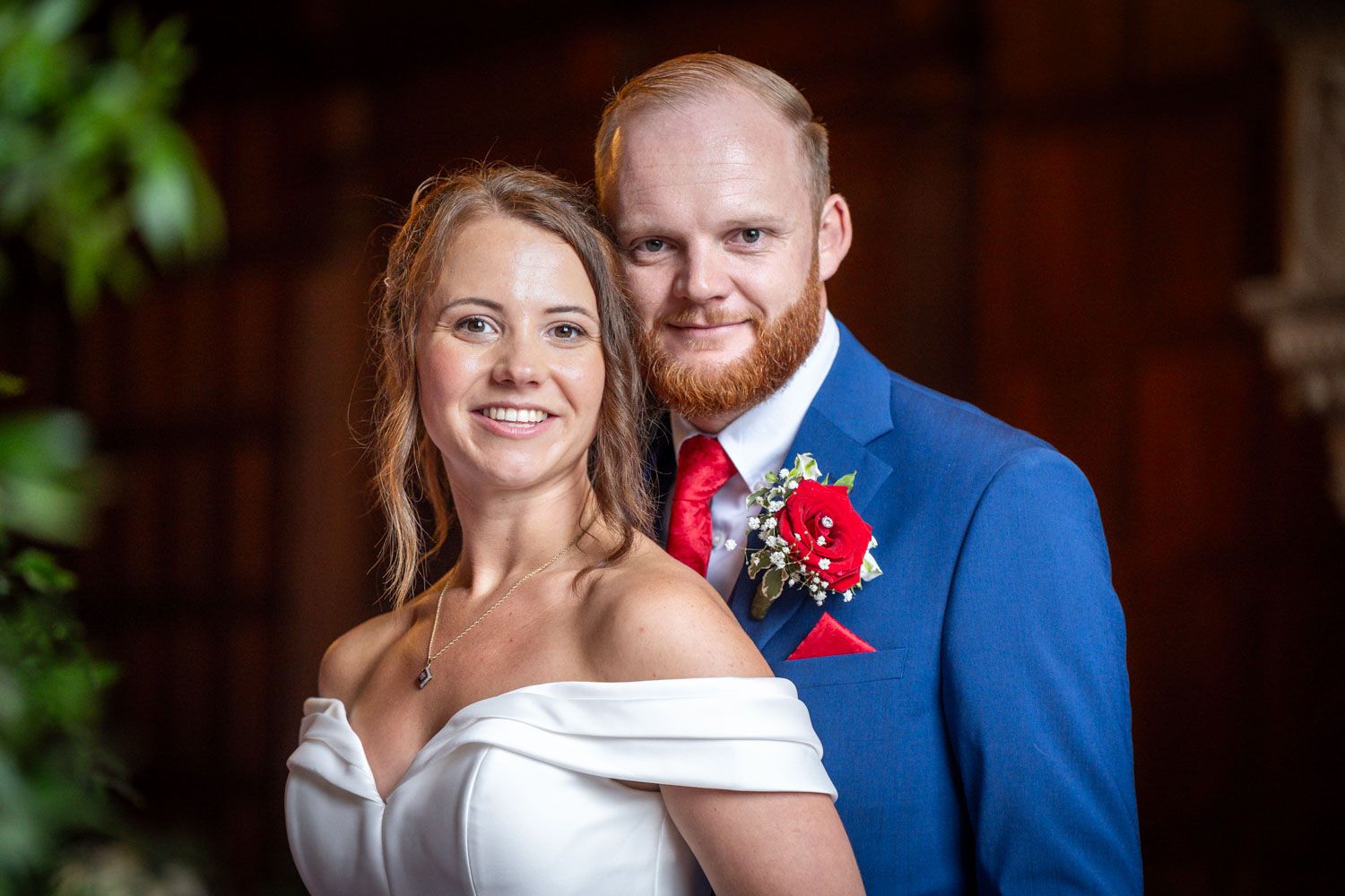 A formal wedding photo showing a couple in a white dress and blue suit with red accents standing together outdoors.