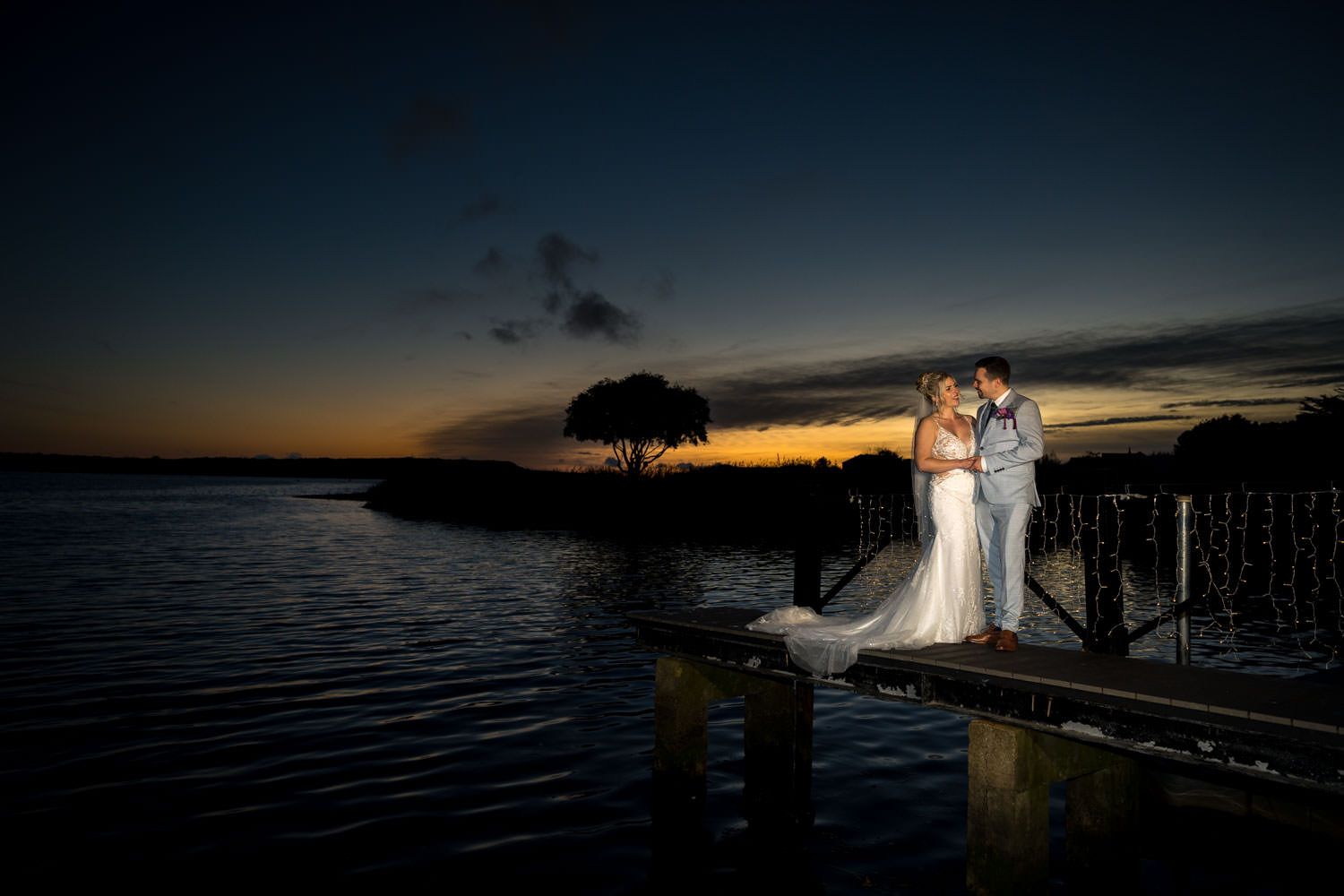 Bride and groom embracing on a wooden dock at stunning golden sunset over calm water.