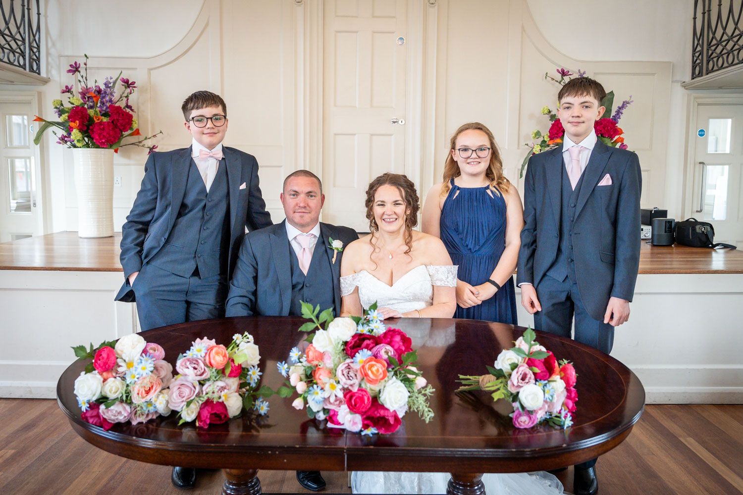 Family portrait during a wedding with floral bouquets of pink and red flowers on an elegant wooden table.