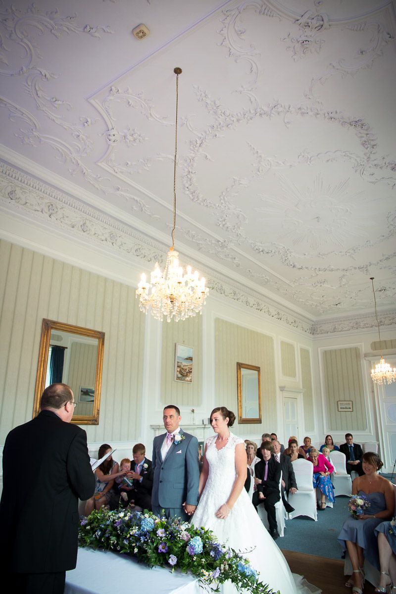 A wedding ceremony takes place in an elegant room with ornate white ceiling details and crystal chandeliers.