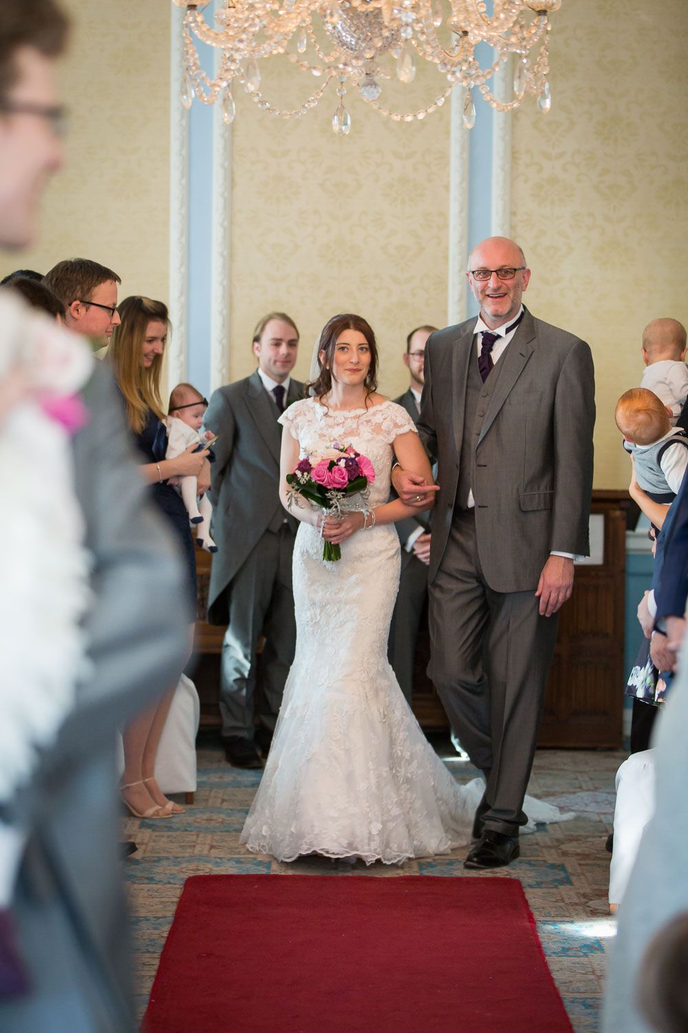 A traditional wedding ceremony taking place in an elegant room with crystal chandelier and red carpet aisle.