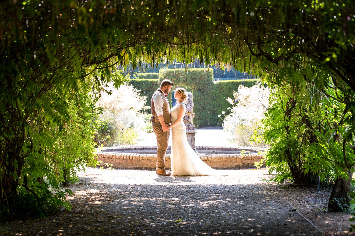 Couple kisses by fountain framed by enchanting green garden archway.