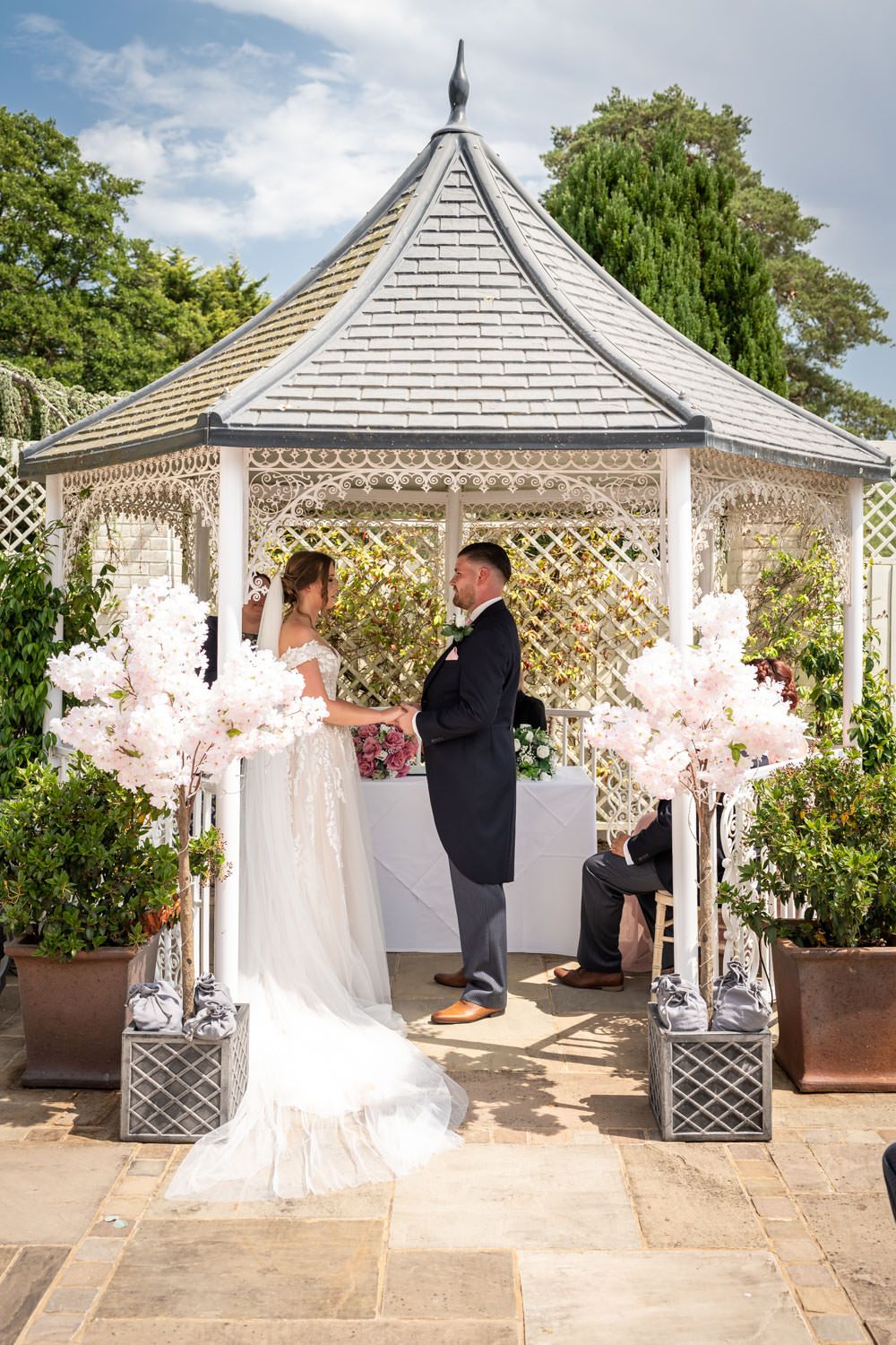 A romantic wedding ceremony takes place in a white garden gazebo decorated with pink floral arrangements.