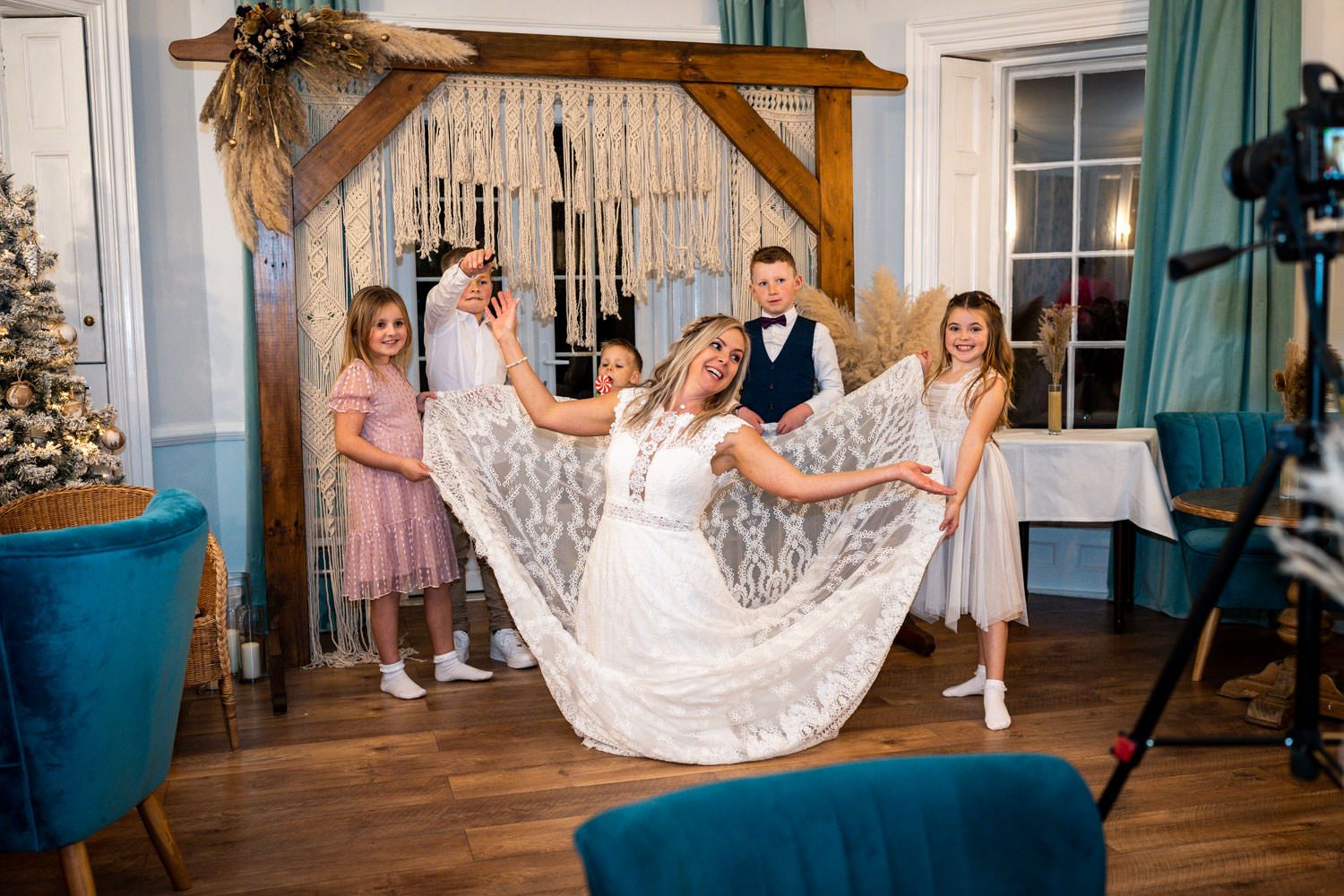 A group stands together in a rustic indoor venue with a macrame backdrop as a lace wedding dress is displayed on hardwood floors.