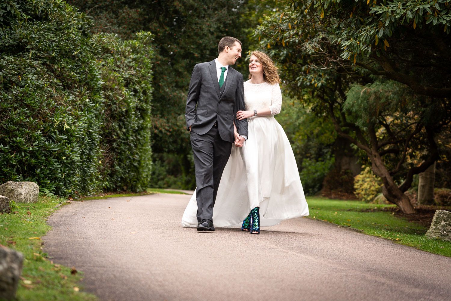A wedding couple walks together on a garden path, with the bride wearing teal shoes beneath her white gown.