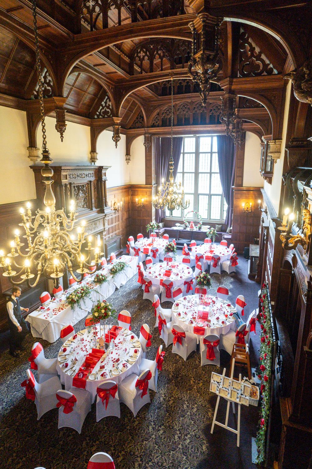 Elegant wedding reception setup in historic mansion with red and white decor, gothic arches and crystal chandeliers.