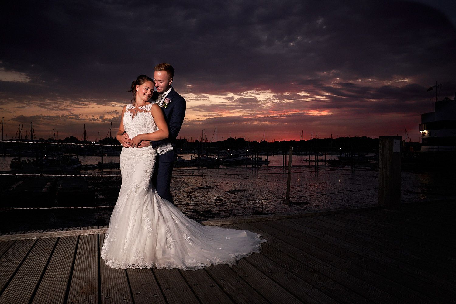 Couple in wedding attire stands on wooden dock at marina during dramatic sunset with boats in background.