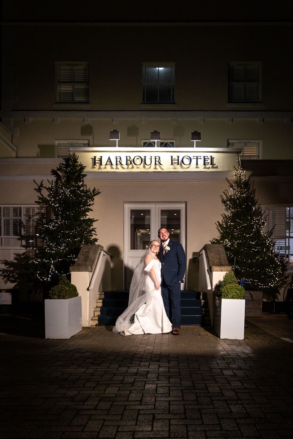 Wedding couple poses for a nighttime photo in front of the illuminated Harbour Hotel with decorated Christmas trees.