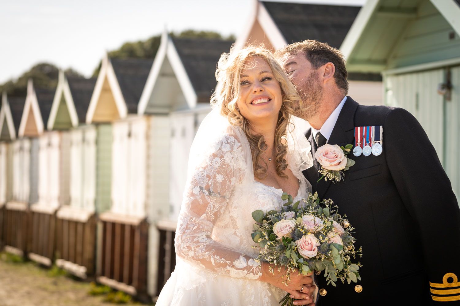 Wedding couple embracing in front of colorful beach huts on sunny day.