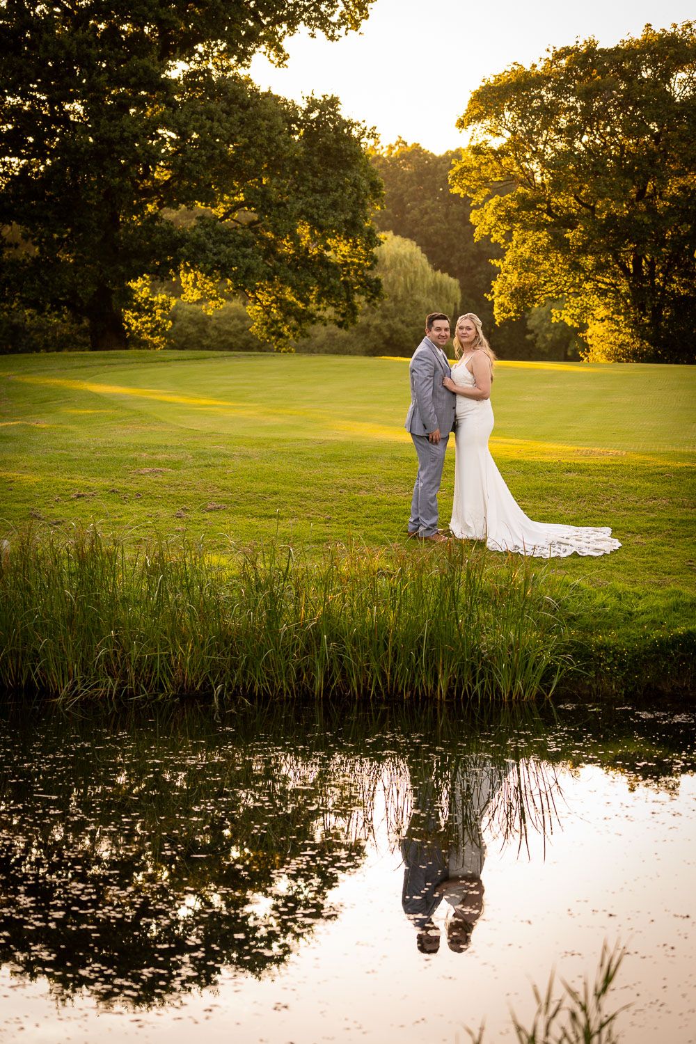 Romantic wedding couple stands beside reflective pond at sunset with golden light filtering through autumn trees.