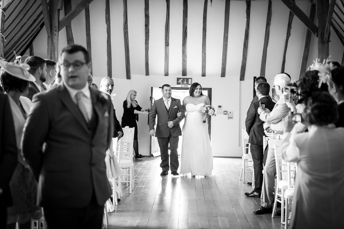 A wedding party walks down the aisle of a rustic barn venue while guests stand on both sides in black and white photo.