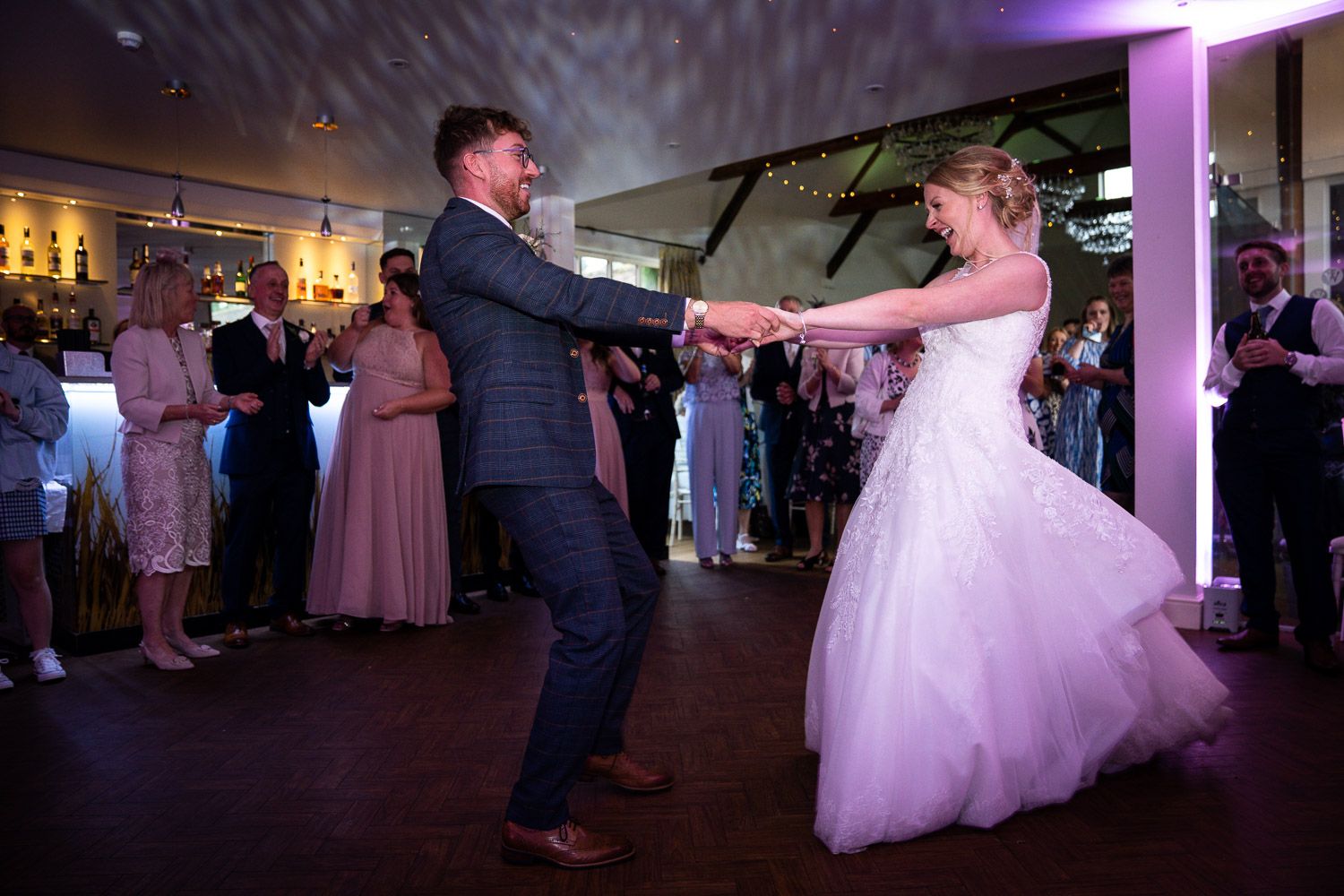 Couple shares their first dance at wedding reception with guests watching from dance floor in dimly lit venue.