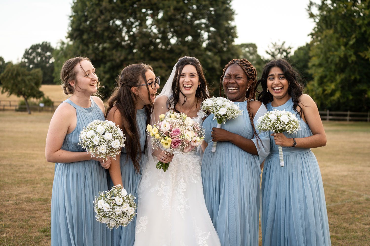 Bride laughing with four bridesmaids in blue dresses holding bouquets outdoors on a sunny day.