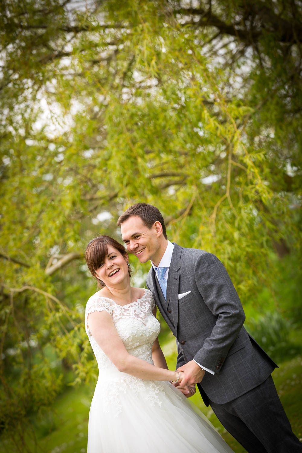 A joyful couple laugh together under green willow trees in their wedding attire.