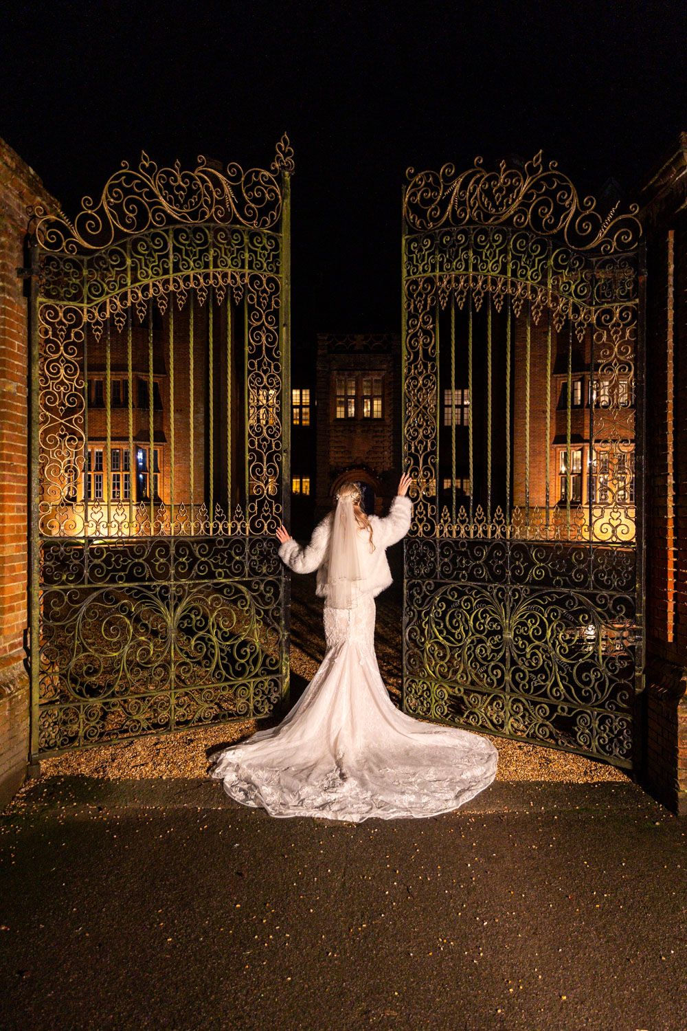 Bride in white gown and veil stands before ornate iron gates illuminated at night.