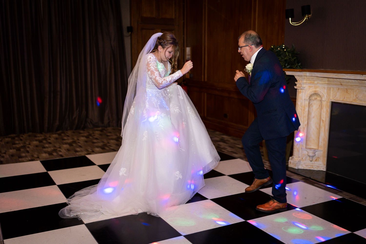 Couple dances on a black and white checkered floor with colorful LED lights reflecting on the bride's flowing white gown.