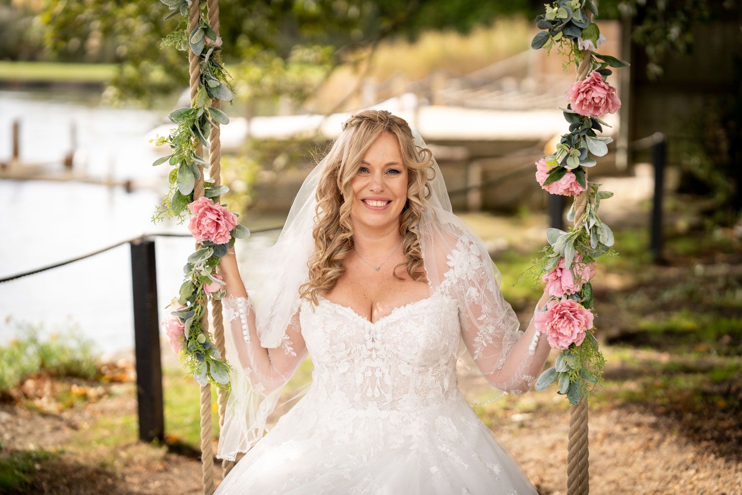 A bride in a white lace wedding dress poses on a floral decorated swing outdoors in a garden setting.