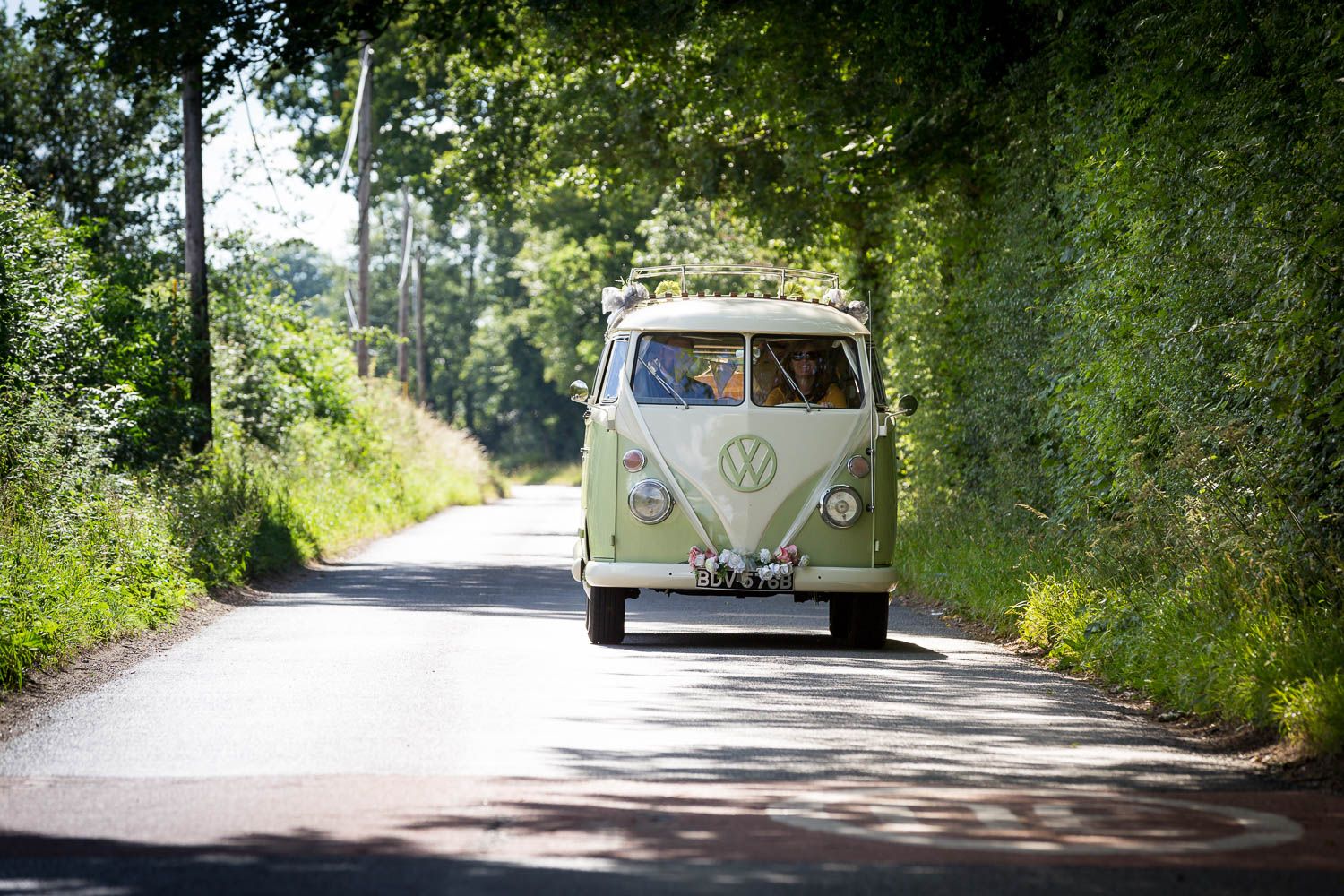 Classic Volkswagen Type 2 bus driving down a scenic country road lined with green trees and foliage.