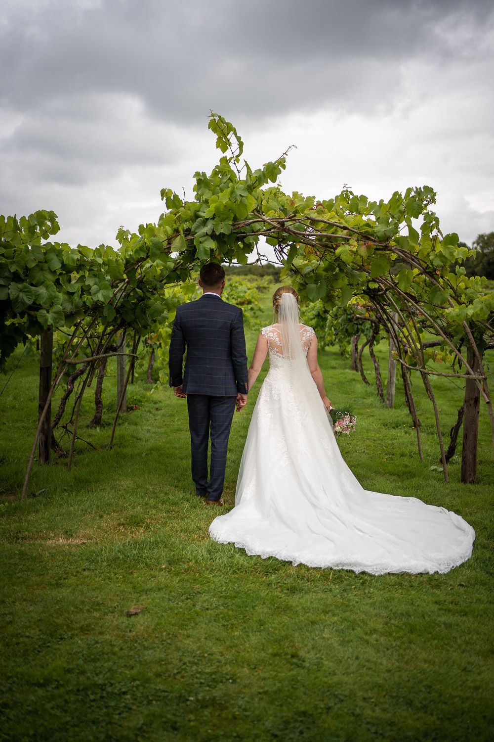 A couple in wedding attire stand together in a vineyard under dramatic cloudy skies.