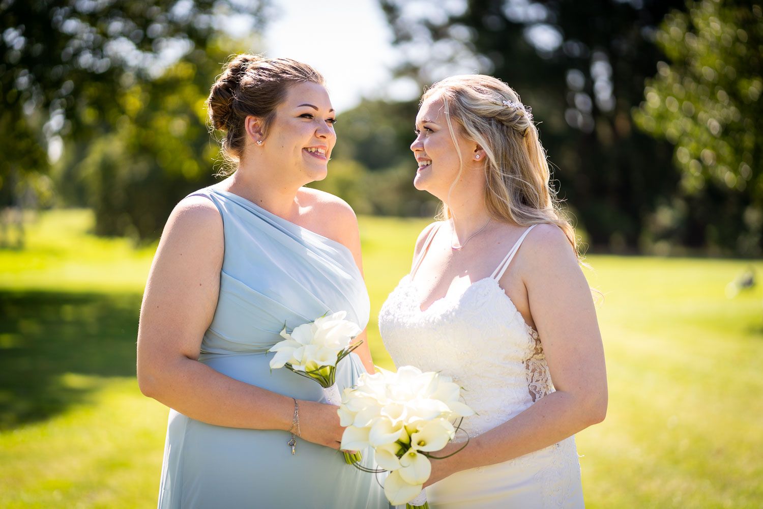 Two people wearing light blue and white dresses share a joyful moment while holding white bouquets in a sunny outdoor setting.