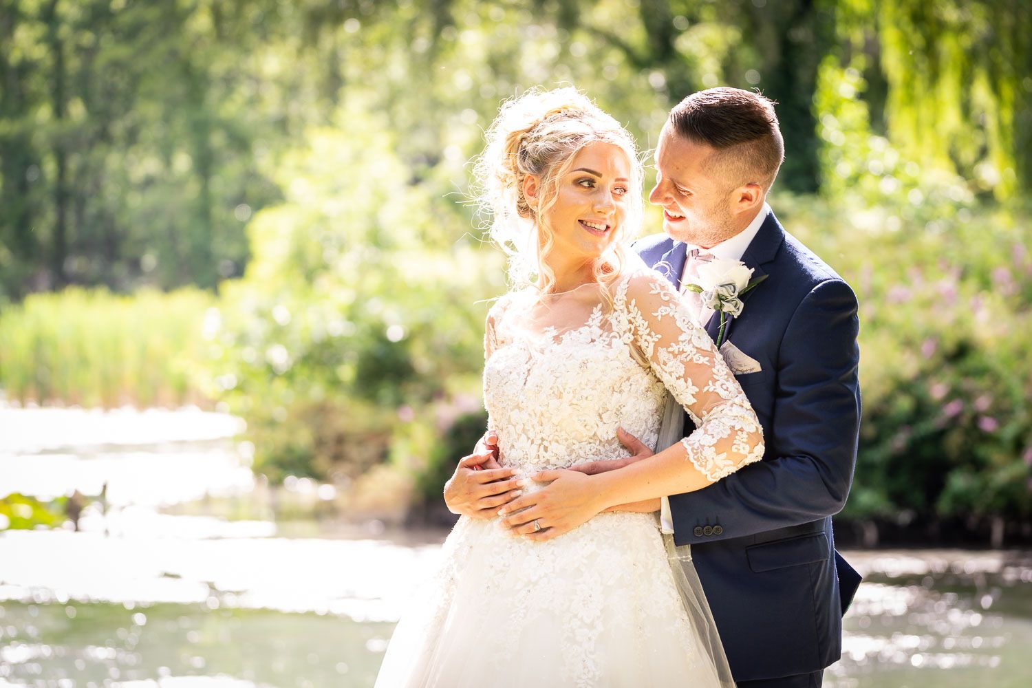 A romantic wedding photoshoot with bride in lace dress and groom in navy suit embracing by a sunlit pond.
