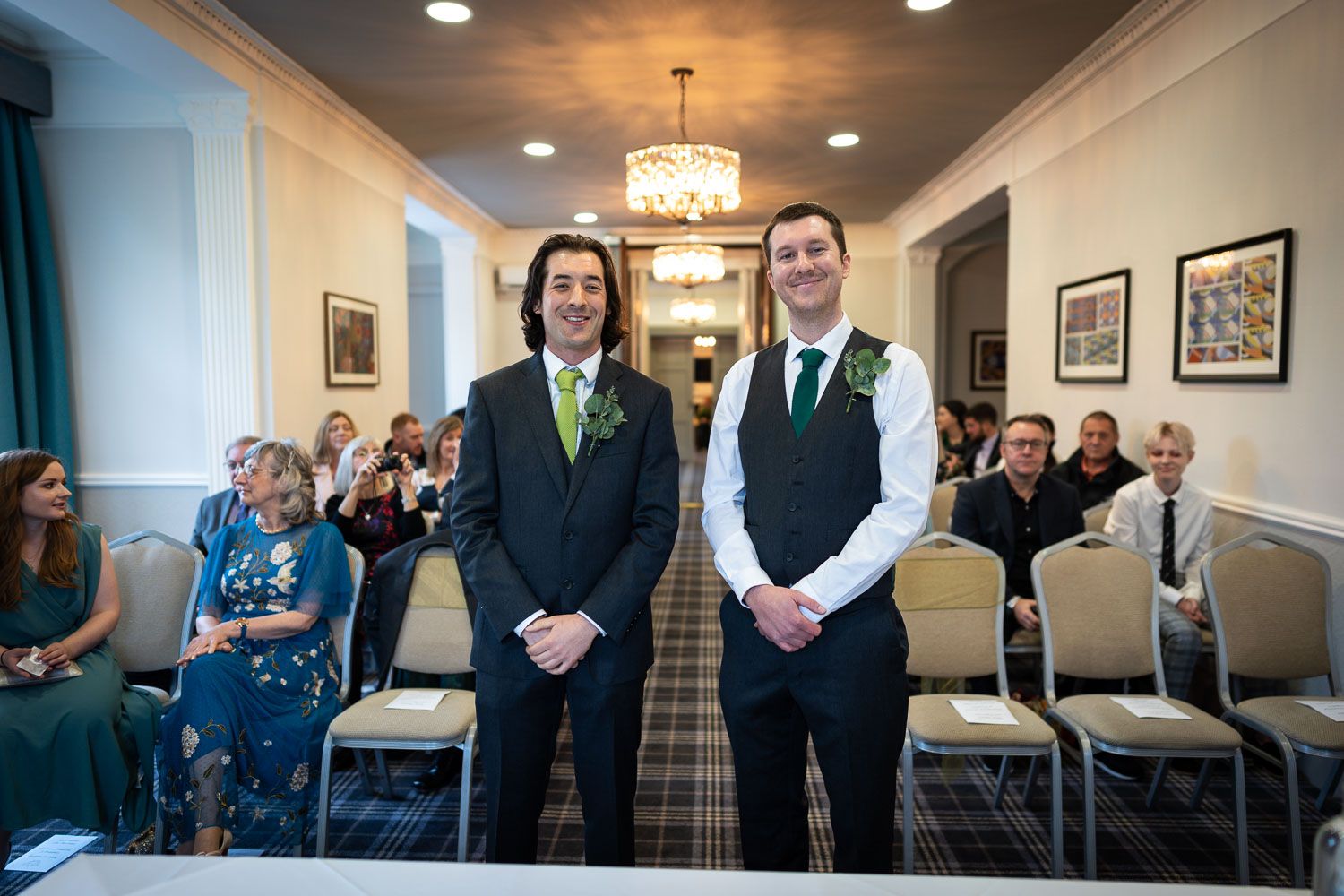 Two men in formal attire stand at the front of a ceremony room with guests seated behind them.