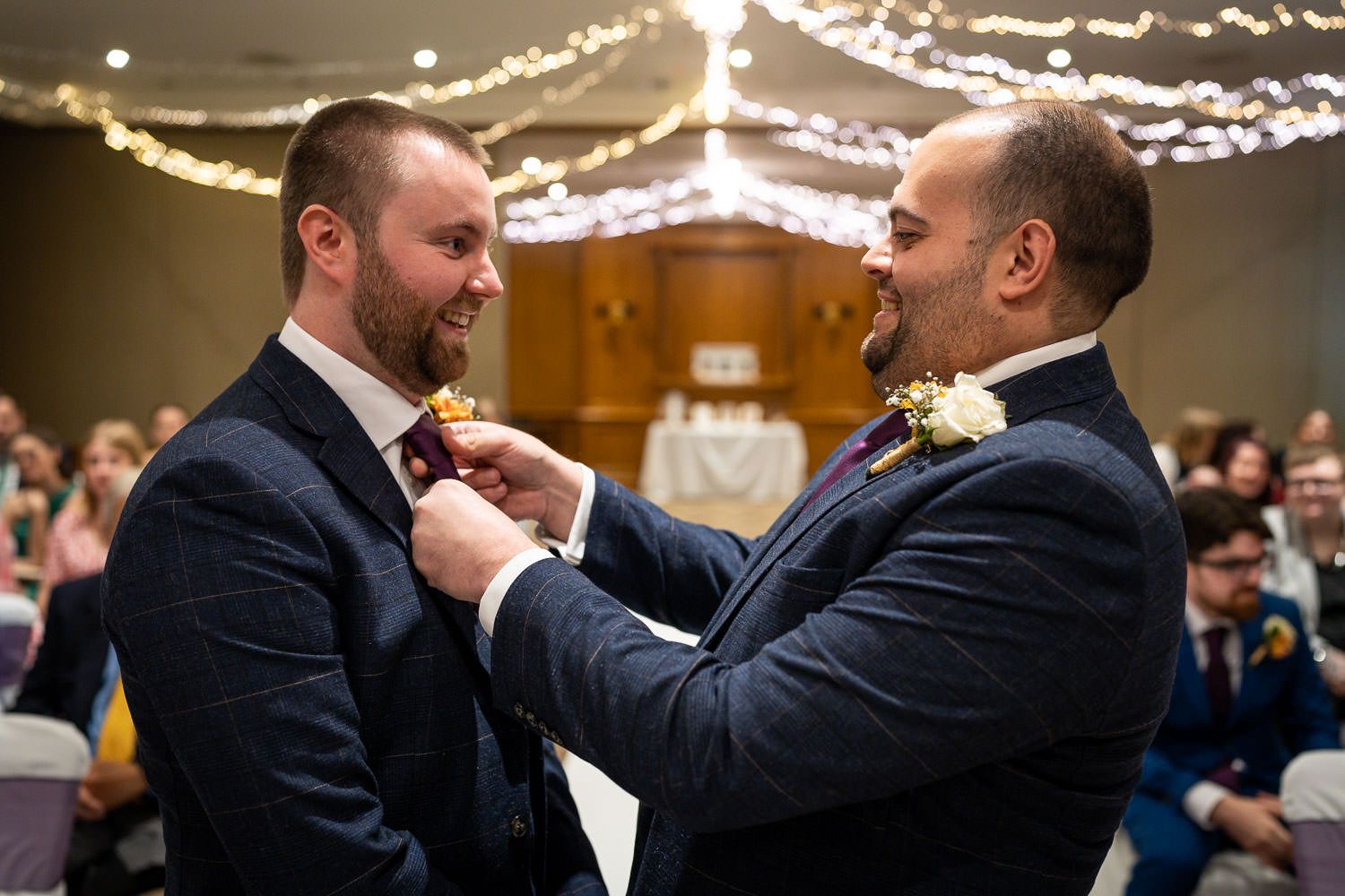 A tender moment captured at a wedding reception as two people in navy suits adjust each other's boutonniere.