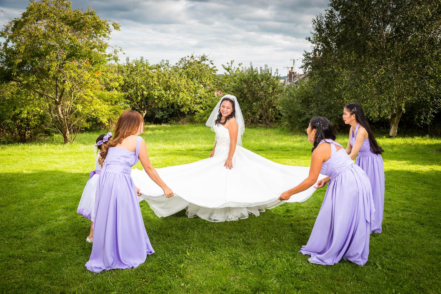 A bride in white surrounded by bridesmaids in purple dresses having fun on a grassy lawn during a sunny day.