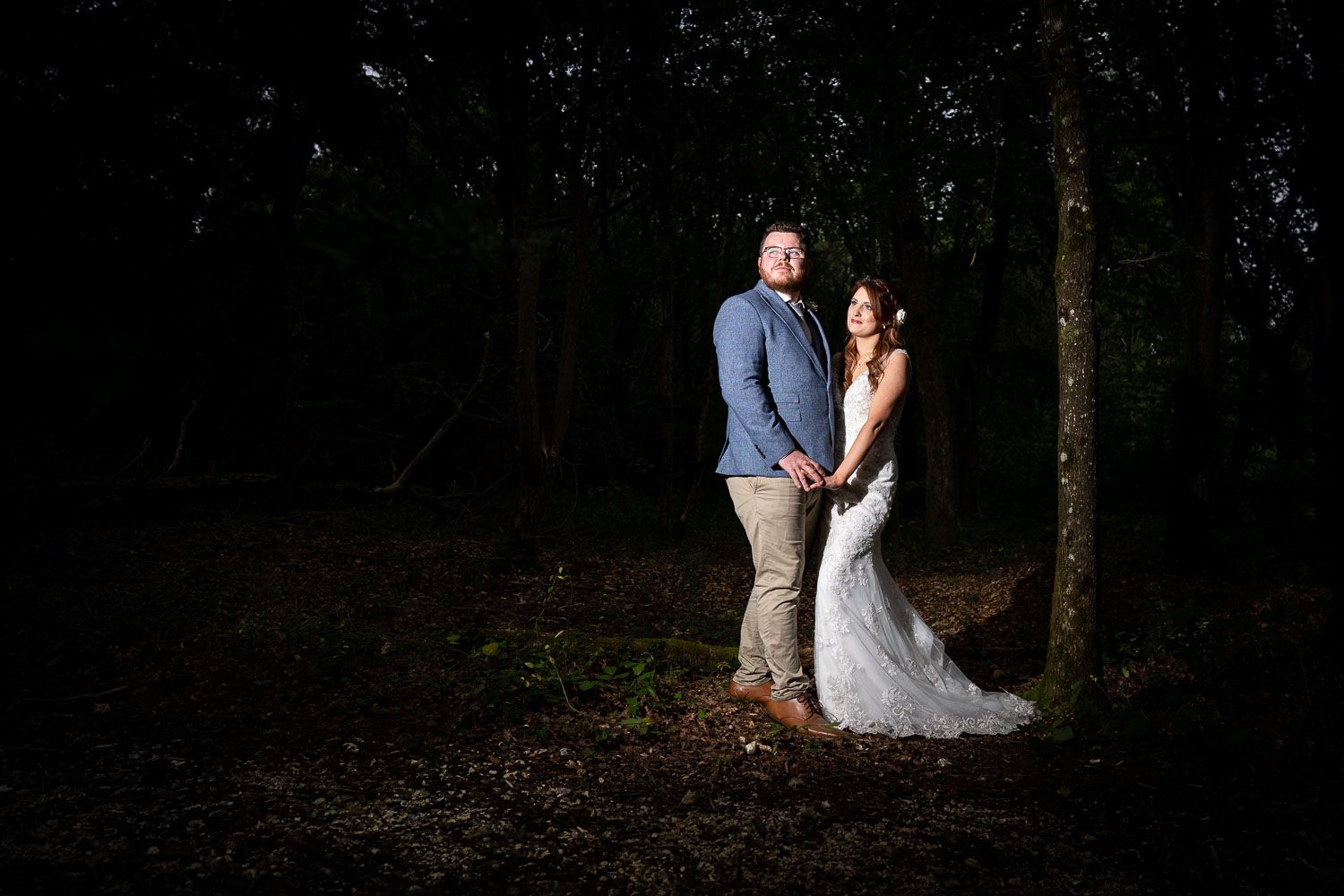 A couple poses in a moody forest setting with dramatic lighting at night, the bride wearing a flowing white dress.