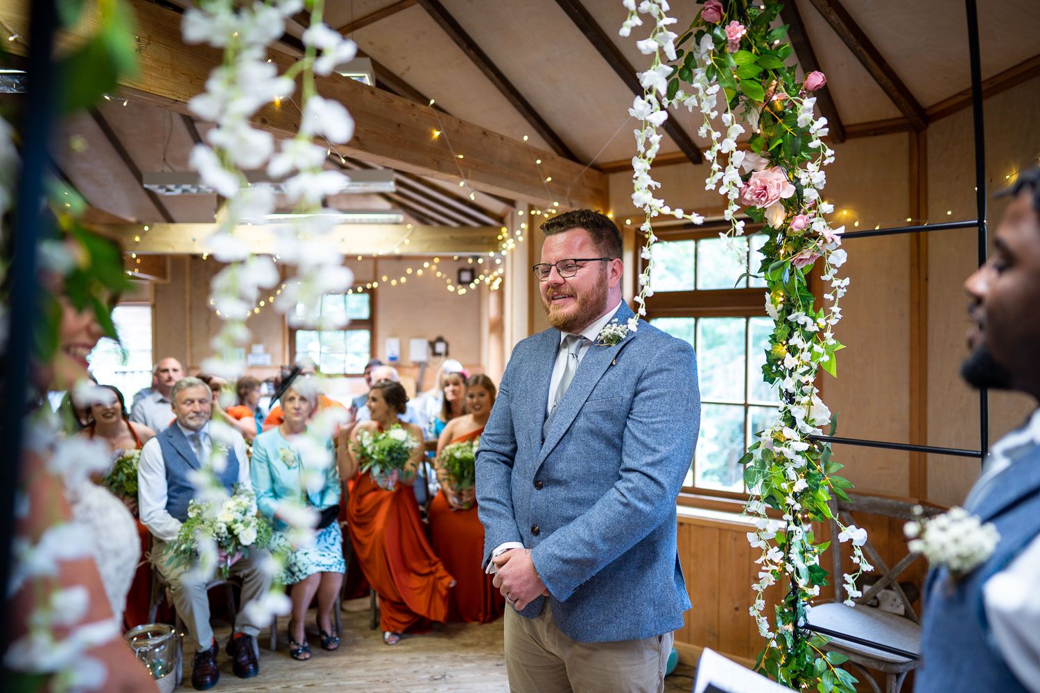 Wedding ceremony at an indoor venue decorated with hanging greenery and floral arrangements as guests look on.