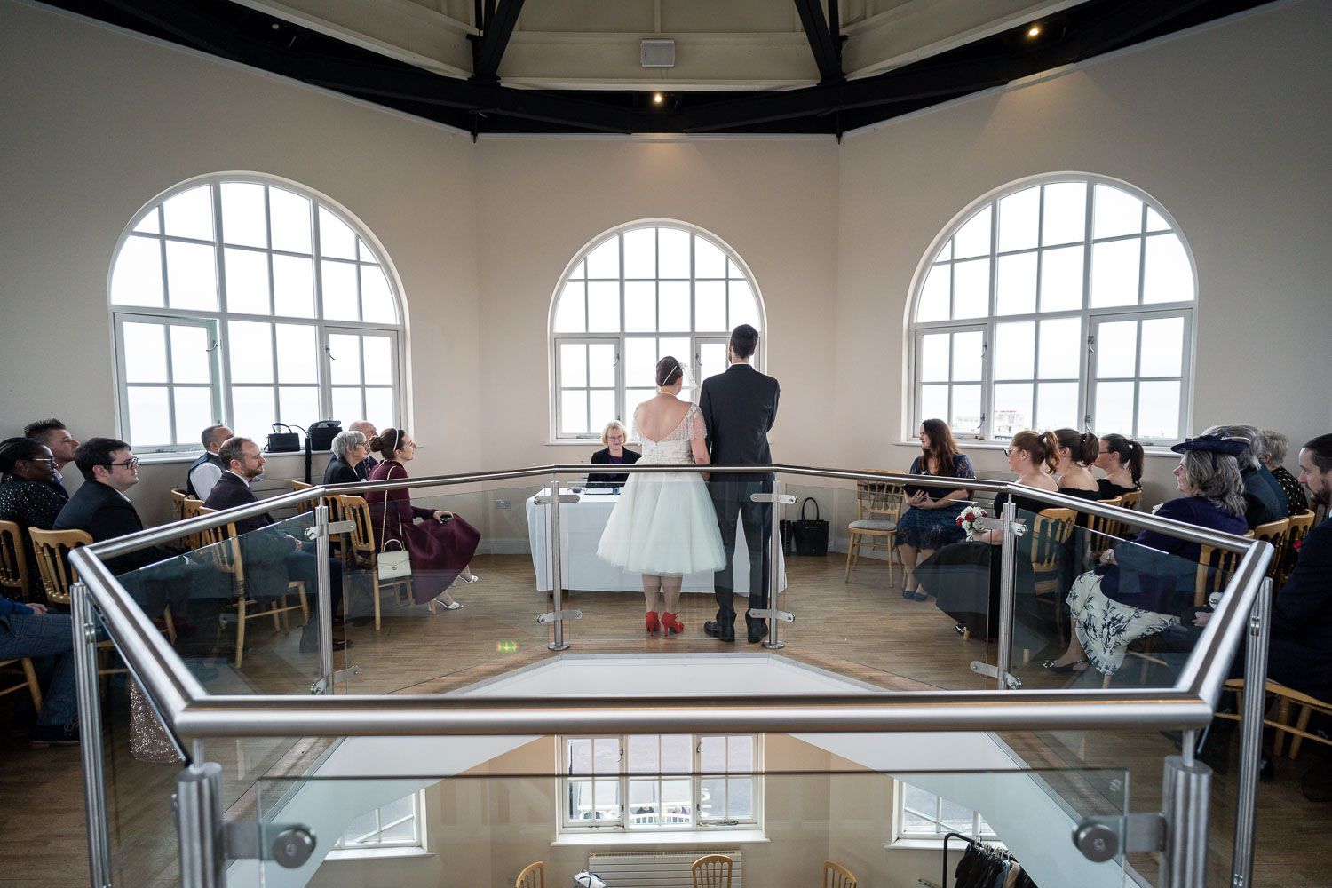 A wedding ceremony takes place in a bright room with arched windows and guests seated in rows along the sides.