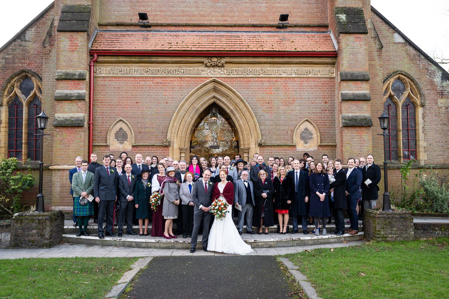 A large group gathers for a formal photograph outside the entrance of a historic brick church with Gothic architecture.