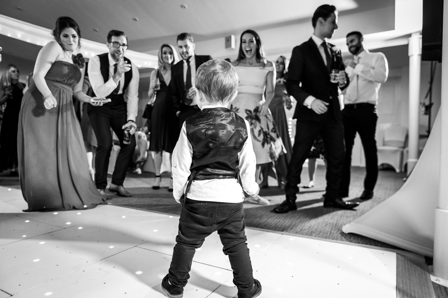 A toddler-aged small child standing on a dance floor during a wedding celebration in black and white.
