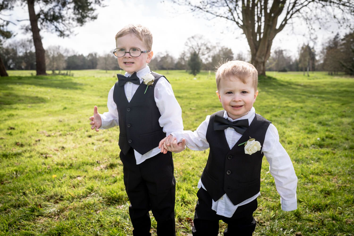 Two young children in matching formal black vests and bowties stand together on green grass in a park setting.