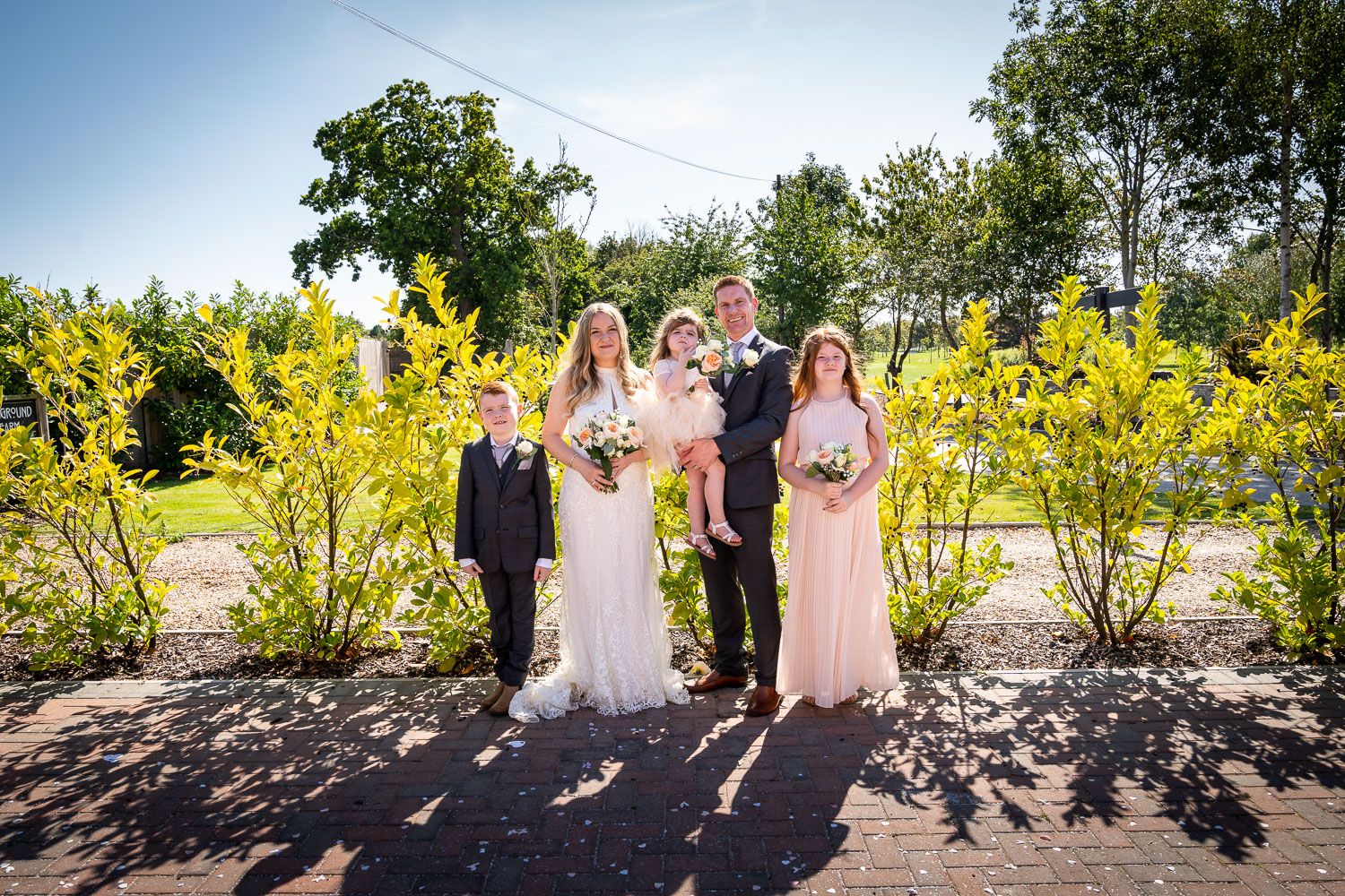 Wedding party stands together on pathway with vibrant yellow forsythia bushes and trees in background on sunny day.