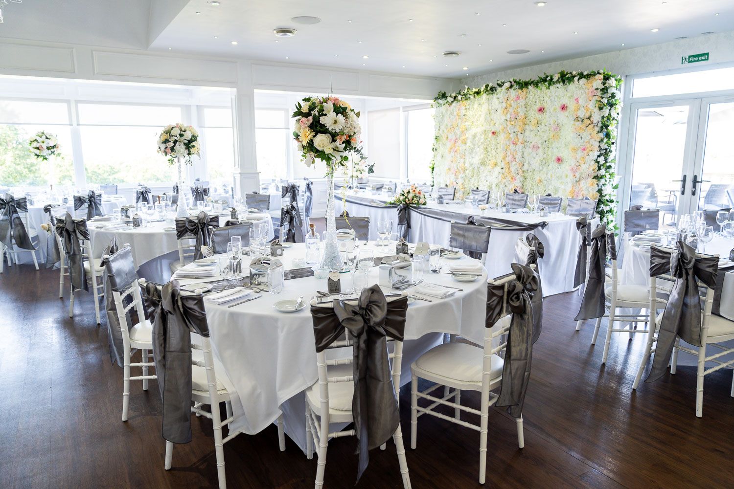 Elegant wedding reception venue with white tablecloths, clear chairs and floral decor against a white flower wall backdrop.