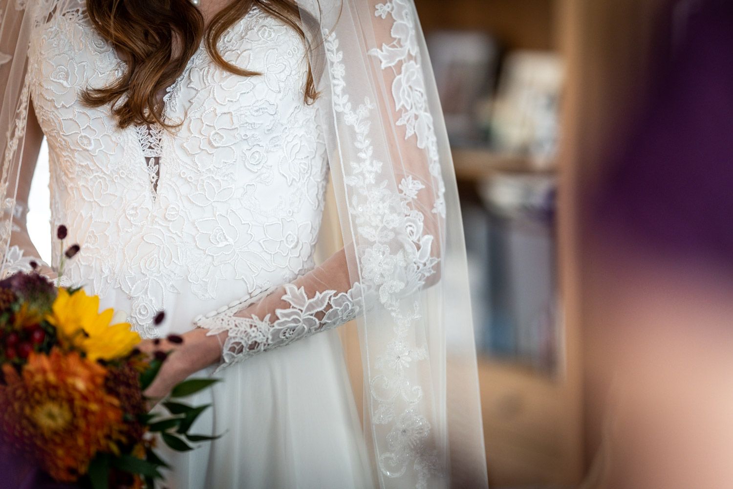 Close-up detail of delicate lace wedding dress sleeve and autumn bouquet with yellow and orange flowers.