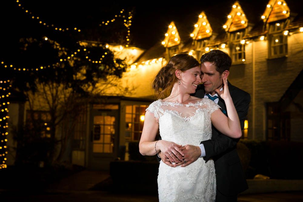 A romantic evening wedding moment with string lights and illuminated buildings glowing warmly in the background.