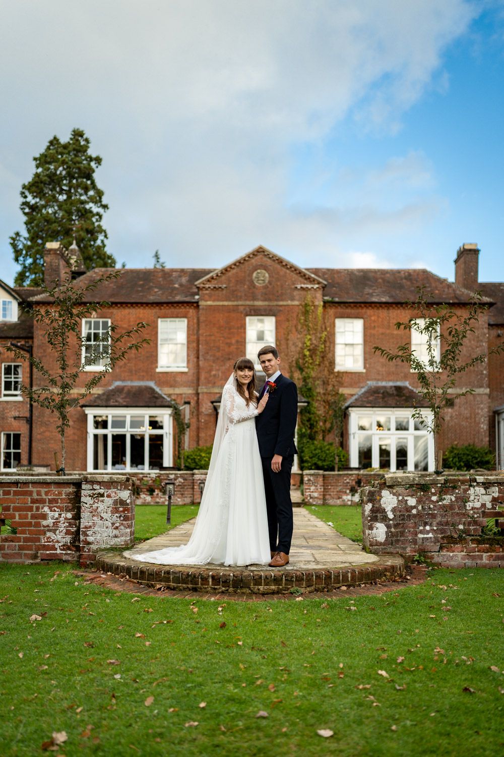 A wedding couple stands on a circular brick platform in front of a grand red brick Georgian manor house with manicured lawn.